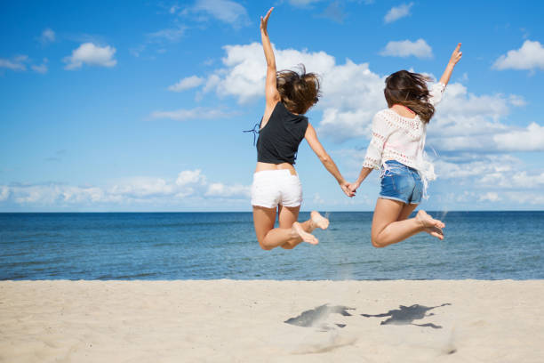 Rear portrait of two female friends jumping on beach holding hands