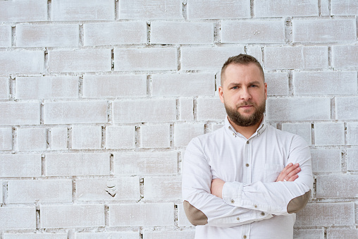 Portrait of serious middle-aged businessman with crossed arms looking at camera and standing near white brick wall
