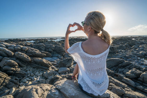 Cheerful young woman on the beach making heart shape frame with hands, beautiful landscape and sunbeams. Freedom travel people relaxation concept. 