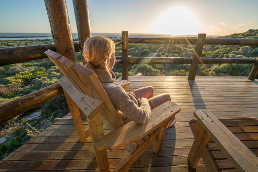 Young woman relaxing on the terrace of her wooden lodge at sunset. Idyllic setting by the sea, shot in the Southern Tip of Africa, Cape Agulhas.