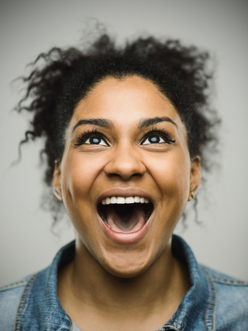 Close-up portrait of a real happy young afro american woman shouting and looking up. Cheerful female is against gray background. Vertical studio photography from a DSLR camera. Sharp focus on eyes.