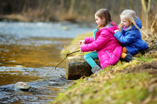 Two cute little sisters playing with a stick by a river on warm spring day