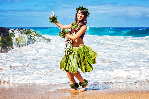 Hawaiian hula dancer in ti leaf skirt, dancing on the beach with ocean waves crashing behind her