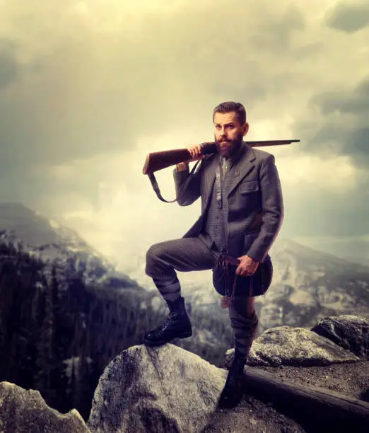Bearded hunter man in old-fashioned hunting clothing with antique gun, rocky mountains and cloudy sky on background. Hunt lifestyle