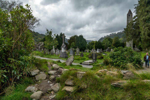 A man walks on a path through an old cemetery in Glendalough, Wicklow County, Ireland. Clouds overhead. Tufts of green grass between the headstones and tumbled rocks.