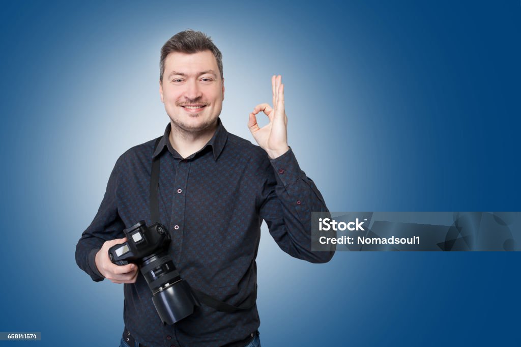 Professional photographer with camera shows ok Professional photographer with digital camera shows ok sign, blue background Camera Operator Stock Photo