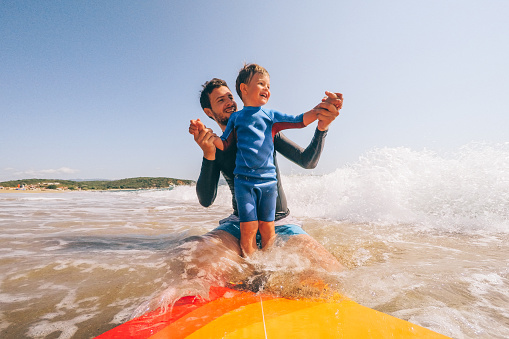 Photo of a cheerful little boy learning to surf on a surfboard with a little help from his dad holding him from behind