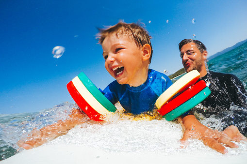 Photo of a cheerful little boy trying to stand up on a surfboard with a little help from his dad holding him from behind