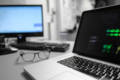 Modern, well-known Laptop computer seen on a desk at a busy office showing both the Laptop running its screen saver together with another type of computer in the background.