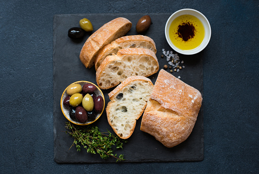 Italian food ingredients background with sliced bread ciabatta, olive oil, olives and rosemary on dark stone slate