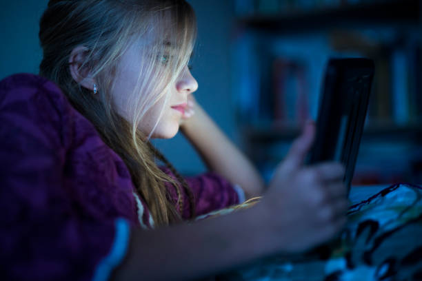 Teenager girl reading from tablet at night in the bed Beautiful long haired teenager girl reading from tablet, in the dark room at night in the bed, with her face illuminated by the screen 13 14 years child teenage girls lying down stock pictures, royalty-free photos & images