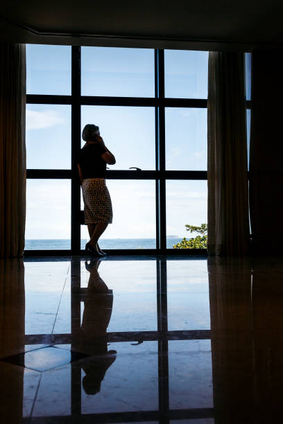 A DSLR Canon photo of a elegant and beautiful senior Caucasian Brazilian woman using her cell phone near a window inside a luxury hotel in Copacabana, Rio de Janeiro, Brazil.