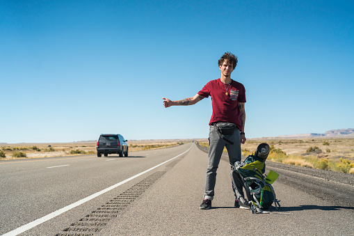 Young man, traveler and hitchhiker, with a big backpack, looking for a ride  on the highway in the desert in Utah, USA