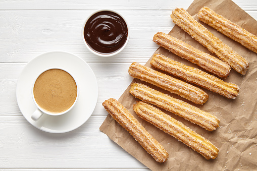 Churros traditional Spain street fast food baked sweet dough snack with chocolate and coffee, rustic decorative parchment paper, white table background. Flat lay top view