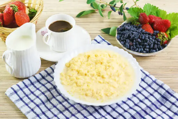 Photo of Corn porridge with milk, a Cup of coffee and the berries of honeysuckle and strawberries.