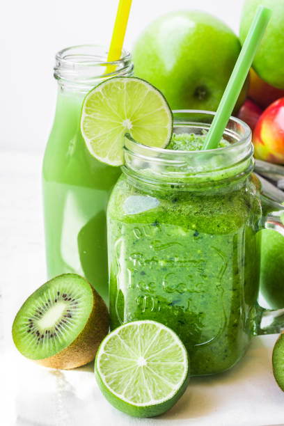 istock Jar mug with green vegetable smoothie, bottle with fruit juice, scattered kiwi, limes, apples, sunlight 658452578