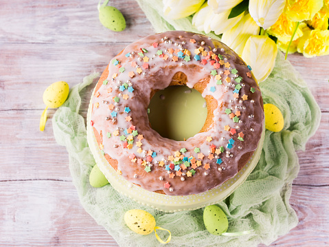 Easter bundt ring cake with sugar frosting sprinkles decorations eggs on wooden table background. Festive holiday home made treat and yellow flowers