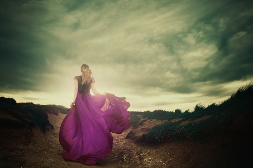 rear view of woman running with her purple flying dress in the wind, enjoying the moment of freedom, dramatic sky.