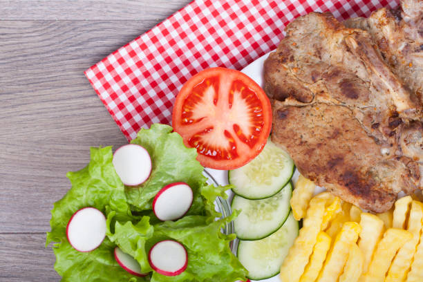 Tasty pork roast on the grill with fries, fresh lettuce, cucumber and tomato on a wooden background. With empty space for your text. View from above.