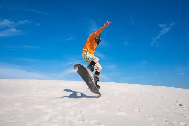 Snowboarder, wearing a scarf to protect against sand, is jumping on a sand dune. Sand boarding in desert. Low point shooting. Wide angle.