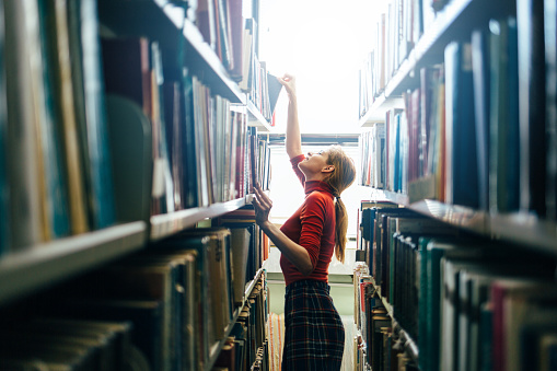 Young librarian searching books and picking one book from library bookshelf.