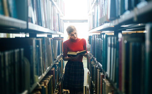 Young woman reading in library between bookshelves.