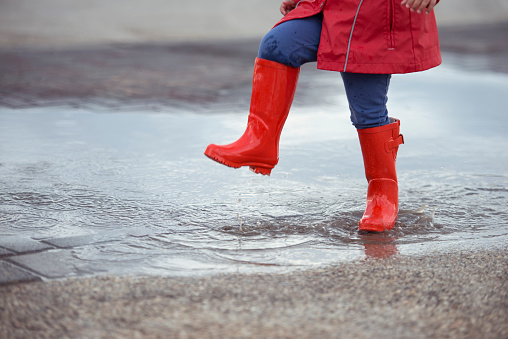 Kid in red rubber boots and raincoat putting his foot into the rain water.
