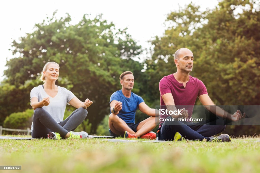 Group of people meditating Group of middle aged people doing yoga sitting on grass. Three people practicing meditation and yoga at park on a bright morning. Mature woman and two mid men meditating together in a lotus position. Yoga Stock Photo