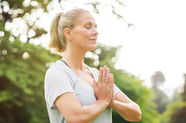 Mature woman in park joining hands with closed eyes and meditates. Relaxed senior woman with joined hands breath deeply with closed eyes. Healthy woman meditating outdoor."r
