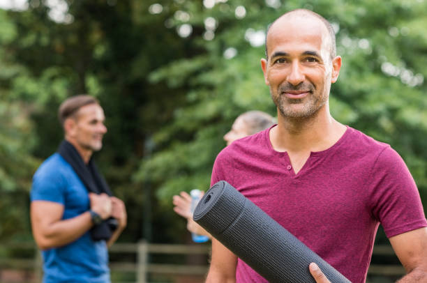 Mature fitness man Smiling mature man holding yoga mat and looking at camera. Portrait of a happy mixed race man with yoga mat at park after fitness exercise. Healthy positive senior man holding yoga mat with people in background. yoga class photos stock pictures, royalty-free photos & images