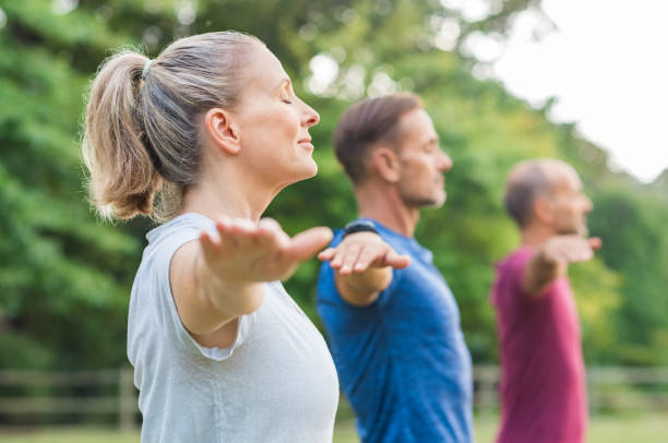 Group of people doing yoga Group of senior people with closed eyes stretching arms at park. Happy mature people doing yoga exercise outdoor on a bright morning. Yoga class with woman and men doing breath exercising with stretched arms. peace park stock pictures, royalty-free photos & images