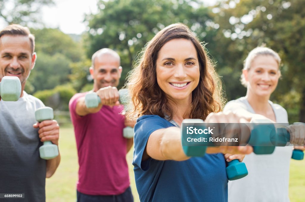 People using dumbbells Mature people in training session of aerobics using dumbbells at park. Happy man and smiling woman practicing fitness together outdoor. Portrait of mature woman doing exercise with other people in background. Exercising Stock Photo