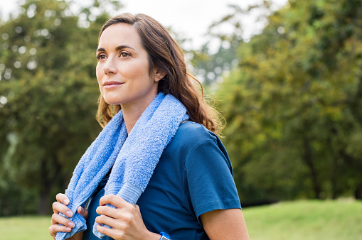 Beautiful fit mature woman resting after workout. Proud brunette woman relaxing with blue towel around neck after yoga in park. Thoughtful mid adult woman thinking after sport and looking away.