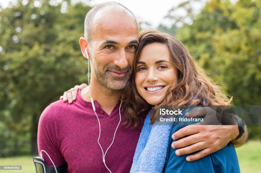 Couple relaxing after jogging Sports couple having fun together at park. Portrait of a happy loving couple relaxing after fitness exercise outdoor. Portrait of multiethnic mature couple embracing and looking at camera. Couple - Relationship Stock Photo