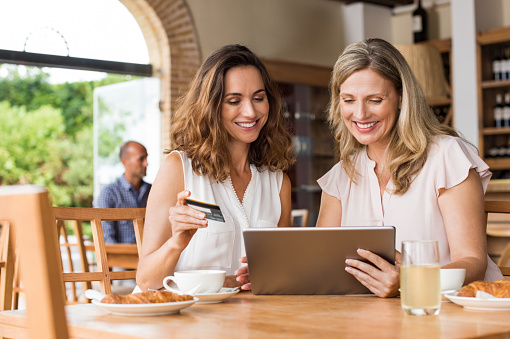 Mature friends buying on internet using a credit card. Happy friends booking and paying their next vacation with debit card on digital tablet in a cafe. Two middle aged women smiling while doing shopping online.