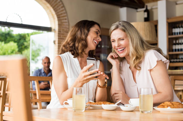 Laughing mature friends Cheerful mature women enjoying a funny video on mobile phone. Mature friends reading a funny message over smartphone. Mid woman showing a cellphone to her friend while laughing over breakfast. women only restaurant stock pictures, royalty-free photos & images
