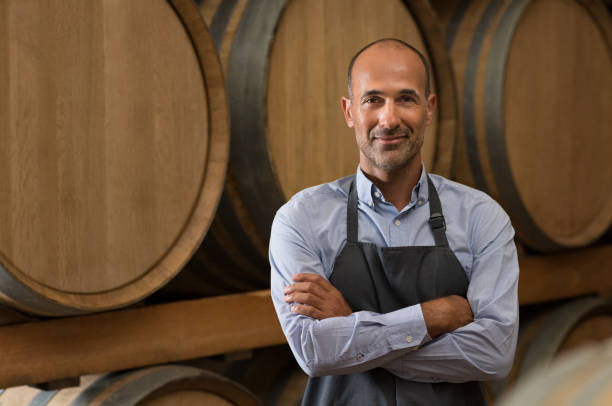 Winemaker in cellar Satisfied winemaker in cellar standing with folded hand. Happy man standing in his warehouse and looking at camera. Smiling professional man standing in wine cellar with wine wooden barrels in background. sommelier photos stock pictures, royalty-free photos & images