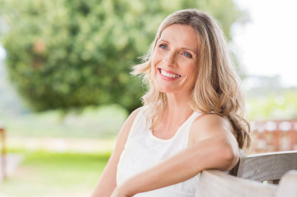 Happy mature woman Happy blonde mature woman sitting outdoor and thinking. Senior woman relaxing on a wooden bench under the tree in a summer day. Portrait of middle aged woman smiling and daydreaming. older blonde woman stock pictures, royalty-free photos & images