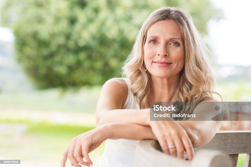 Senior woman relaxing Happy senior woman relaxing on bench in the lawn. Close up face of a mature blonde woman smiling and looking at camera. Retired woman in casuals sitting outdoor in a summer day. Women Stock Photo