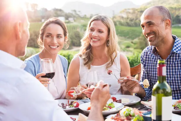 Group of mature people eating together in a vineyard in a summer day. Happy woman sipping wine while talking to friends during a lunch in a winery. Happy senior couple having dinner with wine at sunset.