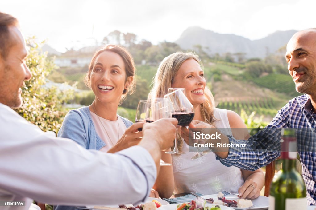Friends raising toast Happy friends raising their glasses in a toast outdoor in a winery farm. Smiling mature woman and men enjoying a picnic together at park. Middle aged multiethnic couple having dinner together and toasting wine."r Friendship Stock Photo