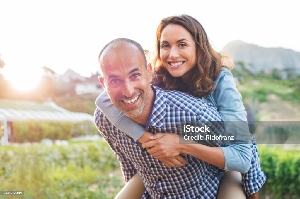 Piggyback mature couple Happy mature couple enjoying outdoors during sunset. Smiling woman piggyback on her man while looking at camera. Portrait of middle aged man carrying on shoulder his wife. Couple - Relationship Stock Photo