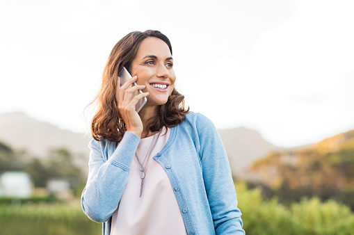 Mature happy woman talking on phone outdoor during sunset. Cheerful hispnic woman using smartphone and looking away. Happy woman in conversation using mobile phone and smiling.