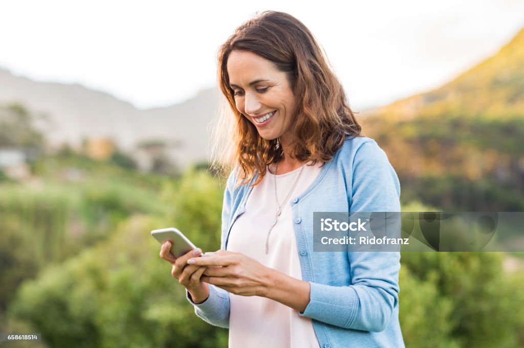 Mature woman using cellphone Mature happy woman using cellphone at park. Smiling woman reading message on smartphone. Brunette latin woman typing a message on her telephone after receiving an email. Women Stock Photo