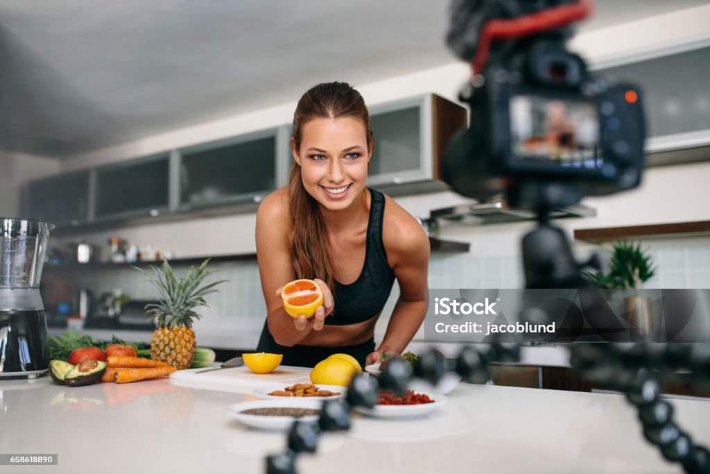 Young female blogger recording content for videoblog in Kitchen. Young woman recording food based video content on camera. Woman showing a cut orange facing the camera. Vlogging Stock Photo