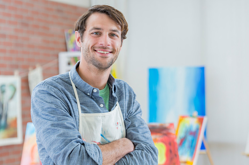 Confident male Caucasian artist stands confidently in his art studio. Paintings are in the background.