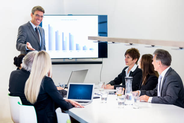 Senior Businessman Business Meeting Financial Presentation Businesspeople together in a team meeting in modern conference room. Senior Businessman showing and presenting the international sales numbers in his presentation on large display. Custom made presentation grafik with different sales areas. Mixed age business group concept shot. cfo stock pictures, royalty-free photos & images