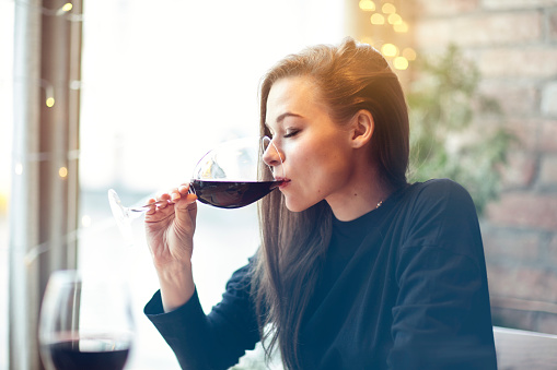 Beautiful young woman drinking red wine with friends in cafe, portrait with wine glass near window. Vocation holidays evening concept