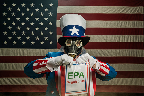 A patriotic Uncle Sam character wearing a full face gas mask while holding a report titled EPA Regulations booklet, with some smoke/smog and a large American flag in the background. Concept of the US government and the Environmental Protection Agency friction under the current Administration.