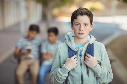 Portrait of sad schoolboy with schoolbag standing in campus at school